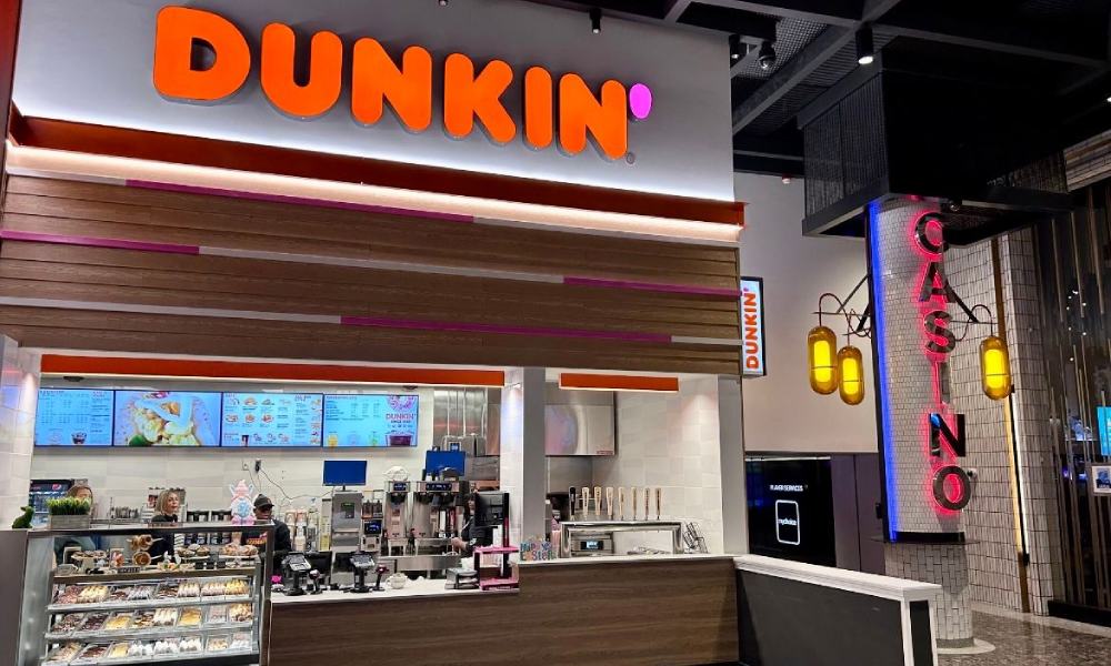 A Dunkin' coffee and donuts franchise kiosk next to a casino entrance. The counter displays baked goods, and the menu is visible above.
