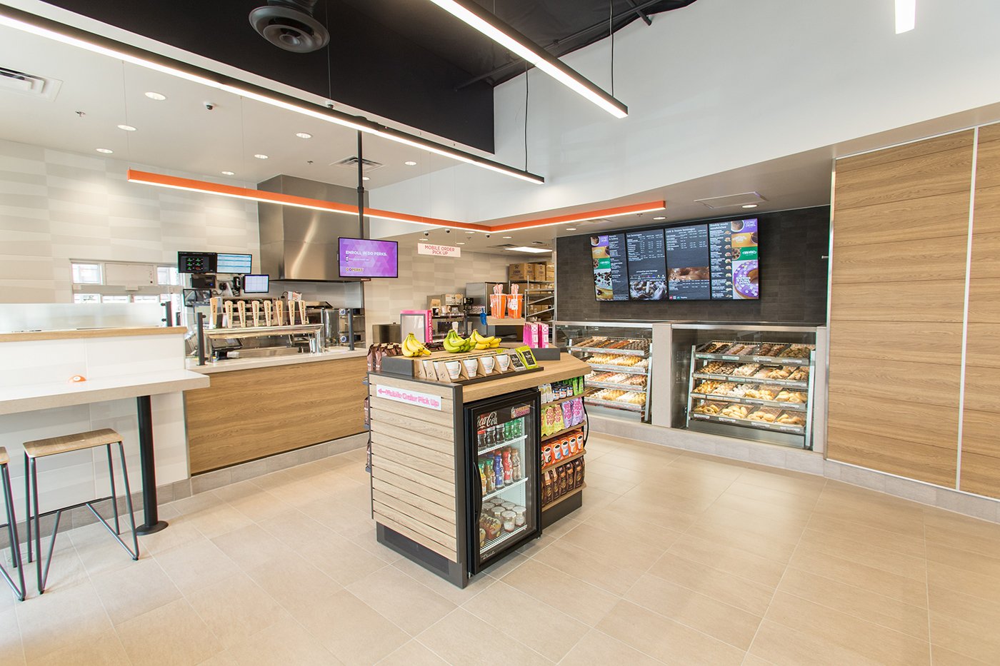 A modern, well-lit coffee and donuts franchise with a counter, digital menu screens, a central island displaying snacks and beverages, and shelves with baked goods. The seating area features high stools and tables.